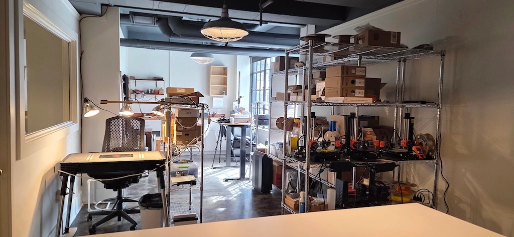 Photo of the 3D Anatomy Studios workshop in March 2026 showing a couple of table workstations and shelving with 3D printers